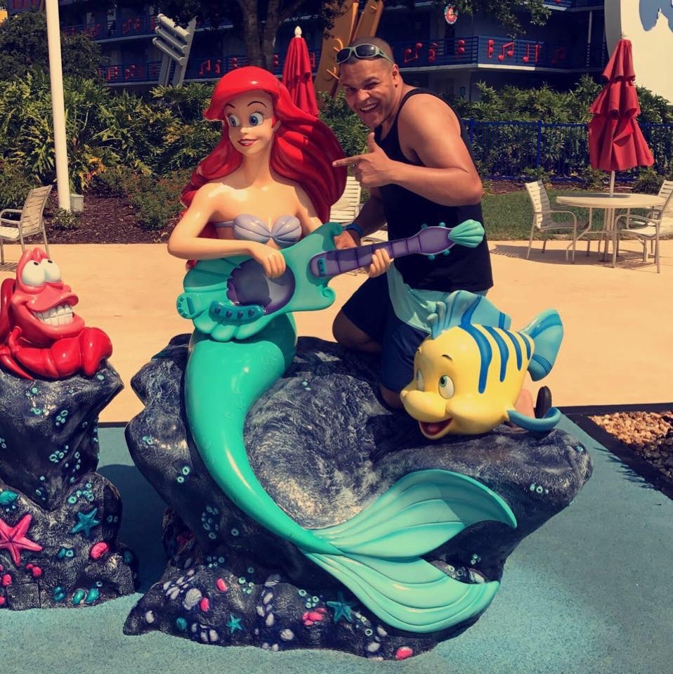 Man in swimming trunks and black tank top posing with Little Mermaid statue at Disney resort pool.