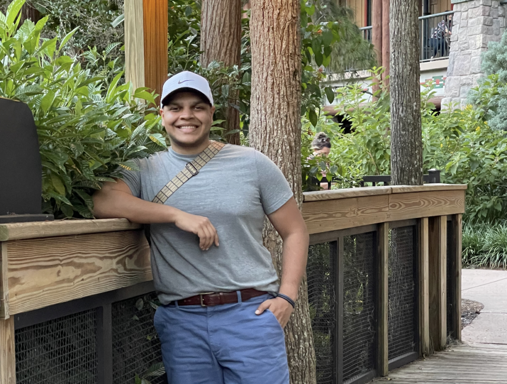 A handsome young man with a bright smile, wearing a grey T-shirt, blue shorts, and a white Nike hat, leans on a wooden railing at Disney's Wilderness Lodge dock, surrounded by beautiful trees and the resort's rustic charm.